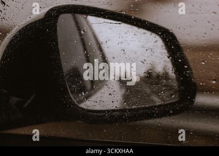 Le gocce di pioggia sullo specchio laterale di un'auto creano una vista suggestiva e soffusa in una giornata di pioggia. Foto ravvicinata dello specchio laterale di un'auto ricoperto da gocce di pioggia e riflessi Foto Stock