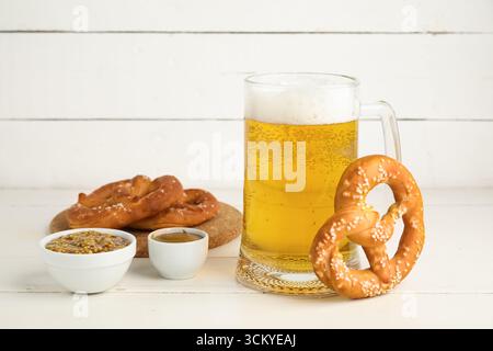 Pretzel, tazza con birra fredda e ciotole con senape su sfondo bianco in legno, primo piano Foto Stock