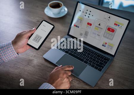 Concetto di interfaccia per app mobili con designer che utilizza smartphone e laptop, mostrando wireframe e layout UX dell'interfaccia utente sullo schermo, simboleggiando lo sviluppo delle applicazioni Foto Stock