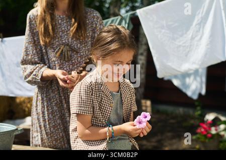 Donna caucasica che intreccia i capelli di una ragazza caucasica sorridente che tiene all'aperto fiori rosa, lavanderia appesa sullo sfondo, volti illuminanti alla luce del sole, interazione familiare informale Foto Stock