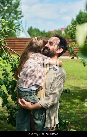 Uomo di mezza età caucasico che tiene e abbraccia la bambina caucasica all'aperto in giardino, sorridente e mostra affetto, alberi verdi ed erba visibili sullo sfondo Foto Stock