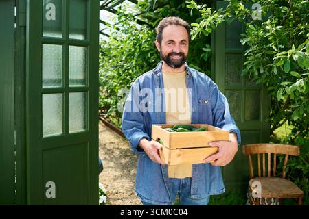 Uomo di mezza età caucasico sorridente mentre trasporta una cassa di legno piena di verdure appena raccolte all'aperto in un giardino, in piedi vicino a porte verdi aperte e piante frondose Foto Stock