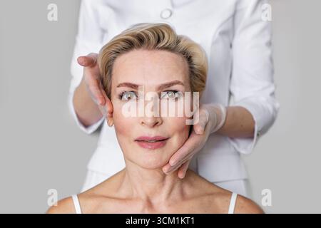 Elegante donna anziana con capelli biondi corti e occhi marroni durante le procedure di cura del viso. Mani cosmetologa che le toccano il viso. Centro di bellezza e anti Foto Stock