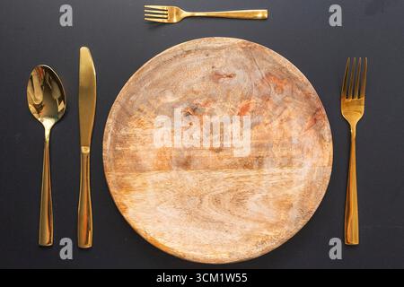 Piatto rotondo in legno con cucchiaio dorato, coltello e forchette disposte su sfondo nero, elegante e lussuoso tavolo per la cena e la presentazione del cibo. Foto Stock