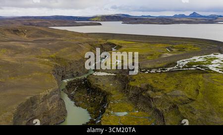 Prospettiva aerea di un canyon in Islanda con un fiume, terreno coperto di muschio, piccole cascate, un grande lago e colline vulcaniche sullo sfondo. Foto Stock