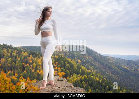 Una bella giovane donna in leggings bianchi posa sulla cima di una montagna. Sport e salute all'aperto. Donna atletica che pratica yoga su una vetta di montagna Foto Stock