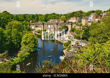 Vista panoramica di un fiume che scorre sotto un ponte ad arco di pietra con una città sullo sfondo, vegetazione lussureggiante e diverse barche a remi sull'acqua a Knaresb Foto Stock