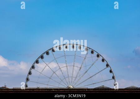 Una grande ruota panoramica bianca con gondole scure è posta su un cielo blu brillante con nuvole sparse, che si innalza sopra una struttura in mattoni a Liverpool, Regno Unito. Foto Stock