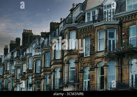 Una fila di vecchi edifici in mattoni a più piani con finestre a bovindo e balconi sotto un cielo nuvoloso, con camini e una luna lontana a Scarborough, Regno Unito Foto Stock
