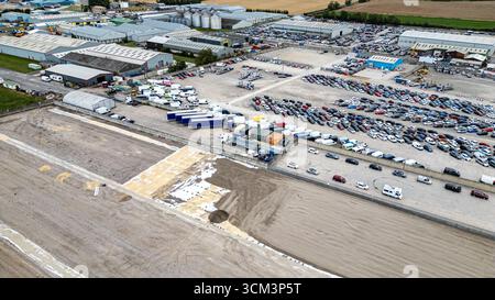 Vista aerea di un grande deposito industriale pieno di numerose automobili, camion e container, adiacente a magazzini e terreni non sviluppati Foto Stock