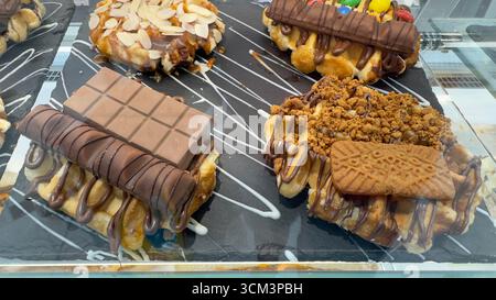 Una vista ravvicinata di quattro deliziosi waffle, ognuno con vari cioccolatini e guarnizioni, presentati su piatti di ardesia nera con gocciolatoi bianchi. Foto Stock