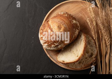 Vista dall'alto di una pagnotta artigianale in pasta madre appena sfornata, presentata su un piatto rustico di legno. Un coltello seghettato poggia accanto al pane, con un mazzuolo Foto Stock
