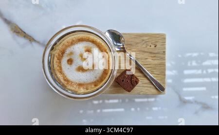 Un caffè di cortado appena fatto con una faccia sorridente servito in una tazza di bicchiere su un tavolino da caffè con un pezzo di brownie. Foto Stock