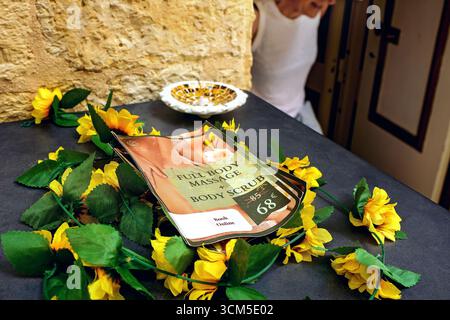 La Valletta, Malta - 08.26.2025: Primo piano di un tavolo con volantino promozionale, fiori gialli e silhouette del visitatore vicino all'ingresso del salone di massaggi. Foto Stock