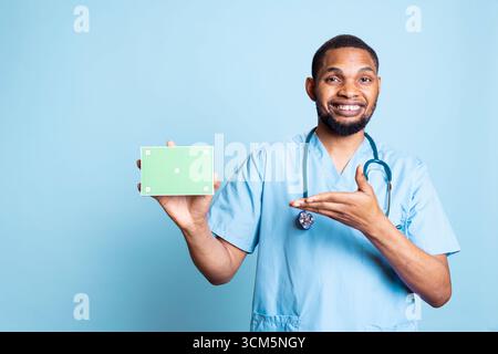 Ritratto di infermiera medica che raccomanda lo schermo verde su una scatola di pillole, sorridendo sulla macchina fotografica e creando una pubblicità. Giovane specialista maschile con scrub e punti stetoscopio nello spazio di copia. Foto Stock