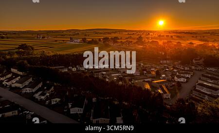 La calda luce del sole tramonta su un tranquillo quartiere di case mobili, evidenziando file di case contro campi verdi e colline lontane. La tranquilla serata Foto Stock