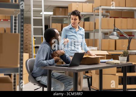 Team di dipendenti multietnici che controlla la logistica di consegna di una piccola azienda, sviluppando un marchio locale. Lo spazio del deposito industriale mostra il coordinamento logistico attivo e il monitoraggio dell'inventario. Foto Stock