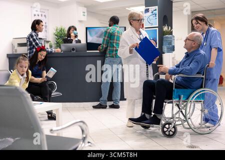 Il vecchio in sedia a rotelle partecipa alla consultazione ospedaliera con le donne professionisti del settore medico. Medico anziano nella hall della clinica, ascolta e offre una guida al trattamento ai pazienti maschi pensionati con disabilità. Foto Stock