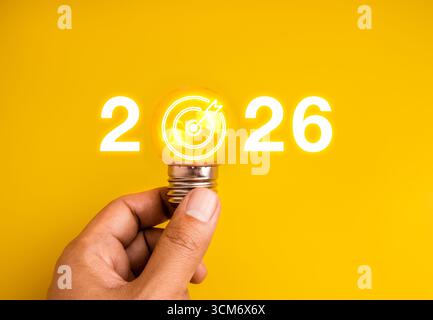 Obiettivo aziendale creativo 2026 con lampadina a incandescenza e icona del tabellone target su sfondo giallo, che simboleggia le idee per il nuovo anno, in Foto Stock