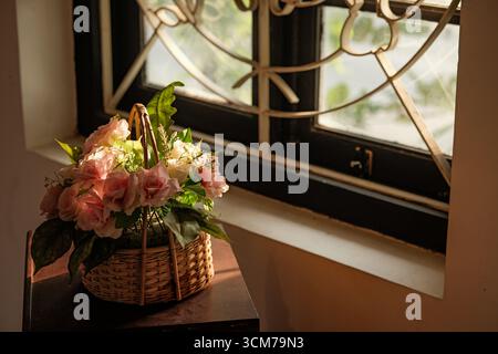 Un cesto decorativo pieno di rose rosa posizionato vicino a una finestra, che illumina la scena con luce solare soffusa Foto Stock