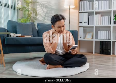 Un giovane uomo siede comodamente sul pavimento con le gambe incrociate, sorridendo mentre utilizza uno smartphone in un salotto accogliente. L'atmosfera rilassata si distingue Foto Stock