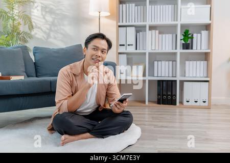 Un giovane uomo siede comodamente sul pavimento con le gambe incrociate, sorridendo mentre utilizza uno smartphone in un salotto accogliente. L'atmosfera rilassata si distingue Foto Stock