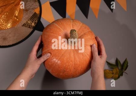 Una persona tiene in mano una grande zucca arancione con entrambe le mani in preparazione all'intaglio. Il cappello da strega e il banner decorativo di Halloween sono sulla superficie grigia. Foto Stock