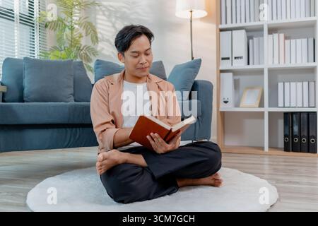 Un giovane è seduto comodamente al chiuso sul pavimento con le gambe incrociate, leggendo un libro con concentrazione ed espressione calma. La scena evidenzia l'apprendimento Foto Stock