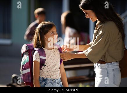La gioiosa riunione si svolge quando la madre abbraccia sua figlia dopo la scuola. Altri bambini giocano e socializzano in sottofondo nelle giornate di sole Foto Stock