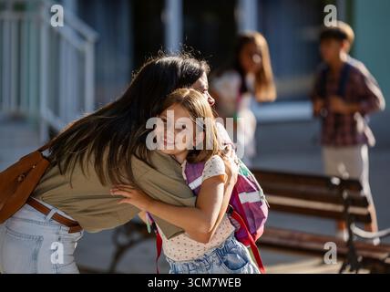La gioiosa riunione ha luogo quando la madre abbraccia sua figlia dopo la scuola. Altri bambini giocano e socializzano in sottofondo nelle giornate di sole Foto Stock