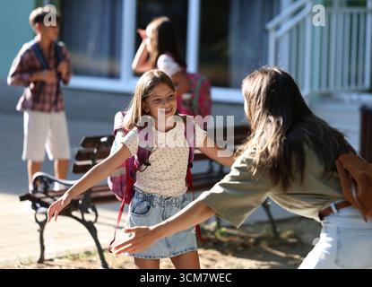 La gioiosa riunione ha luogo quando la madre abbraccia sua figlia dopo la scuola. Altri bambini giocano e socializzano in sottofondo nelle giornate di sole Foto Stock