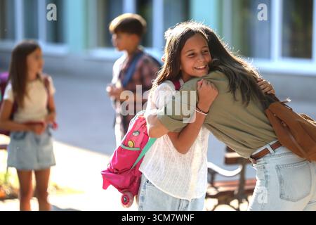 La gioiosa riunione ha luogo quando la madre abbraccia sua figlia dopo la scuola. Altri bambini giocano e socializzano in sottofondo nelle giornate di sole Foto Stock