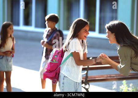 Madre eccitata ha interagito con sua figlia felice, godendosi il loro tempo insieme fuori scuola mentre altri bambini camminano in background Foto Stock