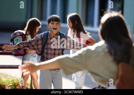 La gioiosa riunione ha luogo quando la madre abbraccia suo figlio dopo la scuola. Altri bambini giocano e socializzano in sottofondo nelle giornate di sole Foto Stock