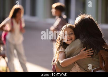 La gioiosa riunione ha luogo quando la madre abbraccia sua figlia dopo la scuola. Altri bambini giocano e socializzano in sottofondo nelle giornate di sole Foto Stock