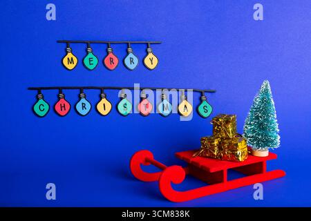 Composizione creativa natalizia caratterizzata da una slitta rossa con scatole regalo dorate, un albero di Natale verde in miniatura e colorate luci a corda che scrivono "M" Foto Stock