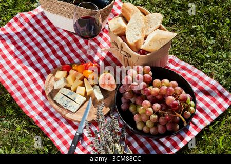 Picnic con formaggio, frutta, pane e vino rosso su una tovaglia a scacchi rossa e bianca. Vista dall'alto. Foto Stock
