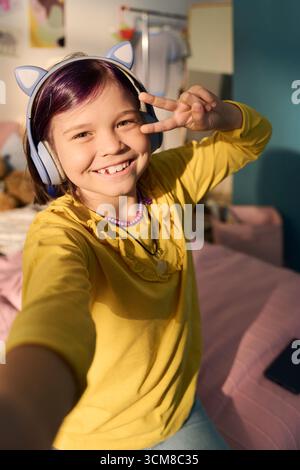 Ritratto di una bambina caucasica sorridente e che fa segno di pace indossando le cuffie con orecchie di gatto e scattando selfie in camera da letto, illuminando il viso alla luce del sole, facendo un'espressione giocosa Foto Stock