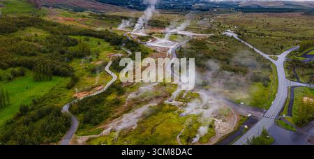 Area geotermica in Islanda con sfiati del vapore in aumento, sentieri in legno, strade asfaltate, terreno verde e marrone e colline e montagne lontane. Foto Stock