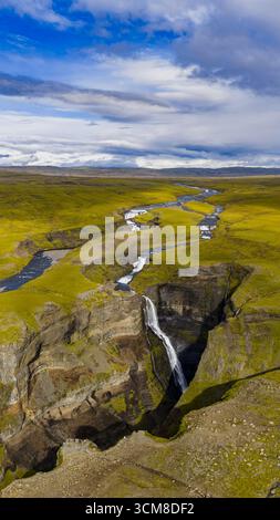 Una potente cascata si tuffa in un profondo canyon, circondato da un terreno coperto di muschio e da un fiume tortuoso sotto un cielo parzialmente nuvoloso in Islanda. Foto Stock