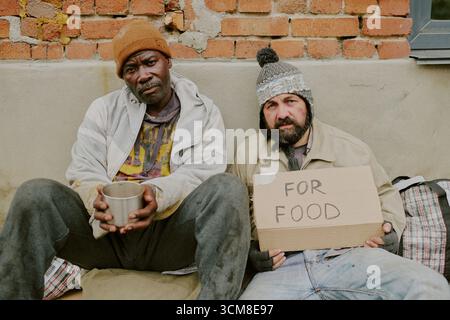 Senzatetto uomo Nero e uomo caucasico seduti all'aperto chiedendo assistenza alimentare Foto Stock