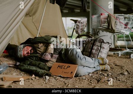 Senzatetto uomo caucasico che dorme all'aperto sotto il ponte tenendo in mano il cartello di aiuto Foto Stock