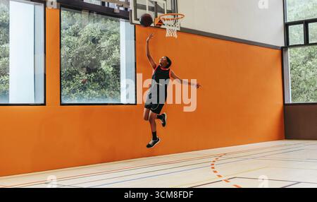 Il giocatore teenager salta in alto per eseguire un pallacanestro in un campo al coperto. Questa immagine cattura l'i, mostrando abilità atletica e passione per lo sport Foto Stock