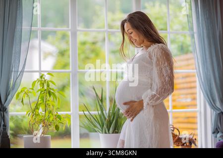 Una donna incinta serena con un vestito bianco sta accanto a una finestra, tenendo delicatamente la sua pancia. La luce soffusa e la vegetazione esterna completano il suo ambiente tranquillo Foto Stock