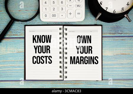 Conoscete i vostri costi. Personalizza il testo dei margini sul blocco note con sveglia, lente d'ingrandimento e calcolatrice su sfondo in legno Foto Stock