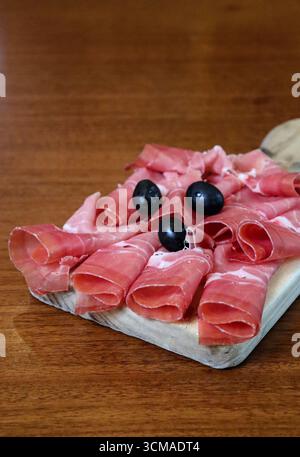 Primo piano di prosciutto tagliato a fette sottili servito su un banco rustico di legno con olive nere. Antipasto tradizionale italiano, salumi di prosciutto, Foto Stock