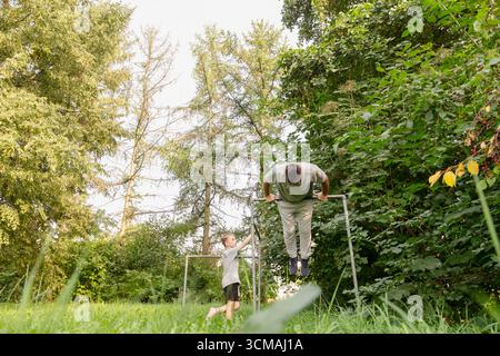 Padre e bambino trascorrono del tempo in un parco giochi circondato da alberi alti e vegetazione. Il bambino guarda mentre il padre esegue degli esercizi Foto Stock