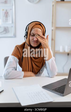 Una giovane donna musulmana seduta alla scrivania con un portatile che le sfrega gli occhi stanchi stressati per il lavoro eccessivo e la stanchezza. copia spazio Foto Stock