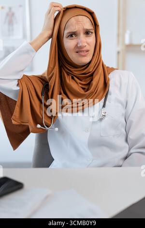 Musulmana medico donna dubita con espressione confuso faccia graffiando testa seduta al tavolo in ufficio clinica. copia spazio Foto Stock