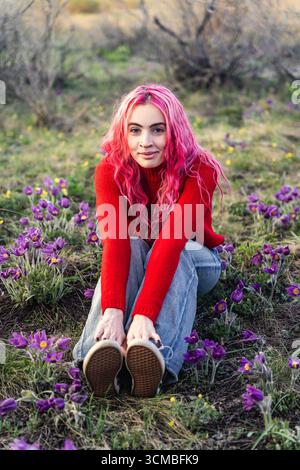 Una giovane donna dai capelli rosa vivaci si siede gioiosamente tra fiori in fiore in un ambiente naturale. Il suo abbigliamento casual e la sua espressione brillante riflettono un sens Foto Stock